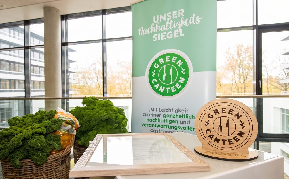 GreenCanteen Siegelfahne, Zertifikat und hölzernes Siegelschild auf einem weißen Podium im Betriebsrestaurant