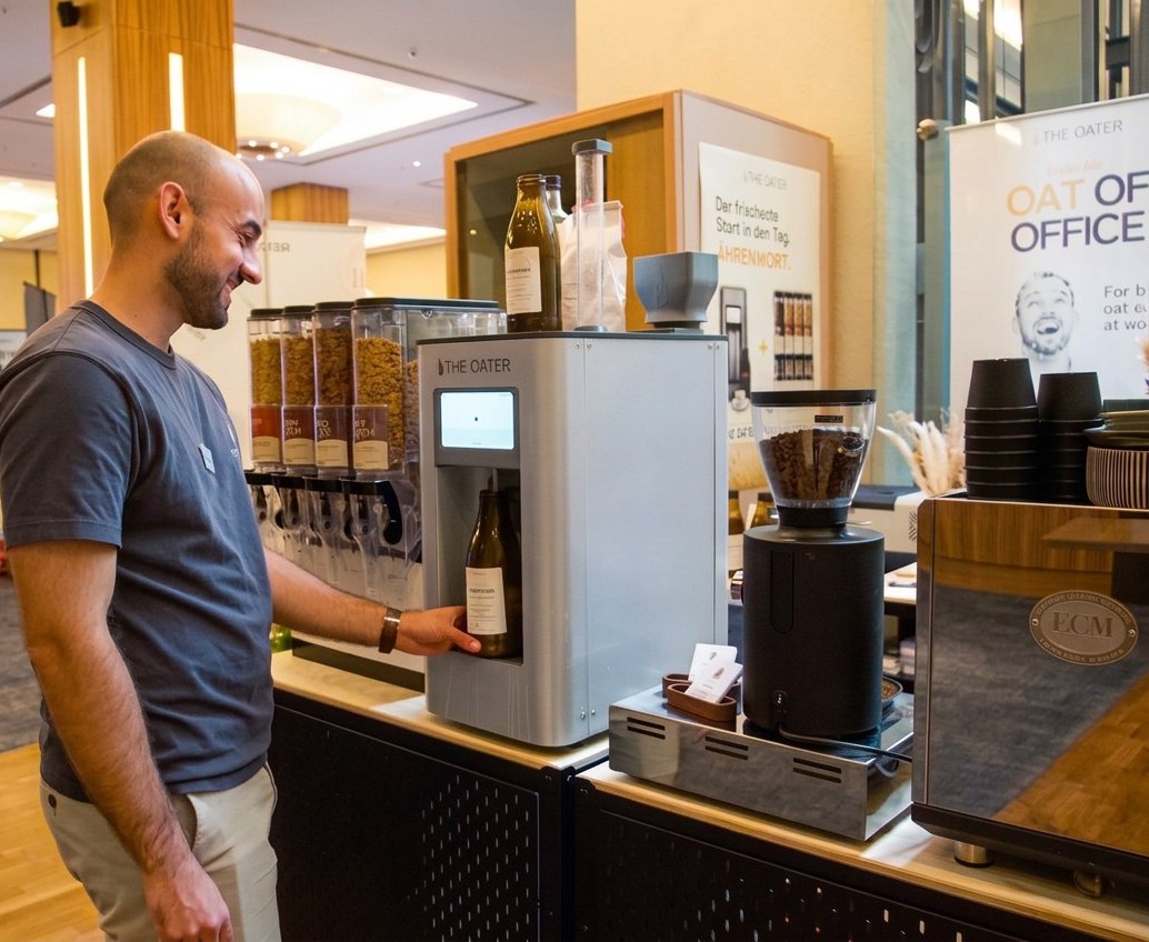 Mitarbeiter entnimmt eine Flasche frischen Haferdrink an der The Oater Maschine neben Müslispendern und Kaffeemühle