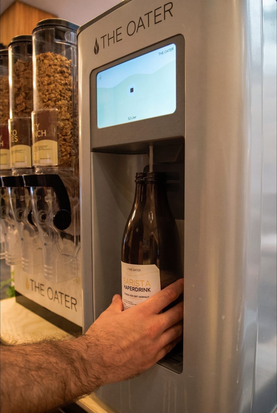 Eine Hand füllt frischen Barista Haferdrink an der The Oater Station in eine Glasflasche, daneben ein Müslispender mit Granola