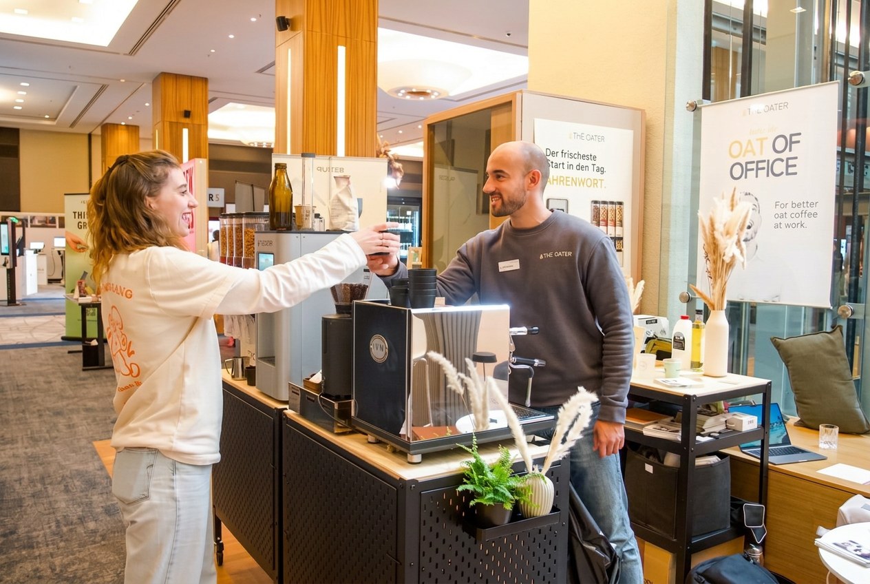 Eine Besucherin und ein Barista stoßen an der The Oater Kaffeebar lächelnd mit ihren Kaffeebechern an