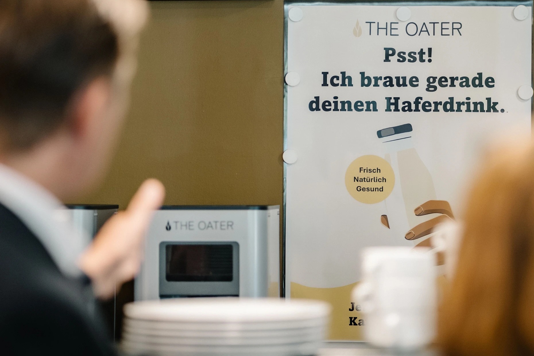 The-Oater-Maschine auf einem Tisch vor einer olivgrünen Wand mit weißem Plakat mit der Aufschrift Psst! Ich braue gerade deinen Haferdrink, davor unscharf Teller, Tassen und Personen