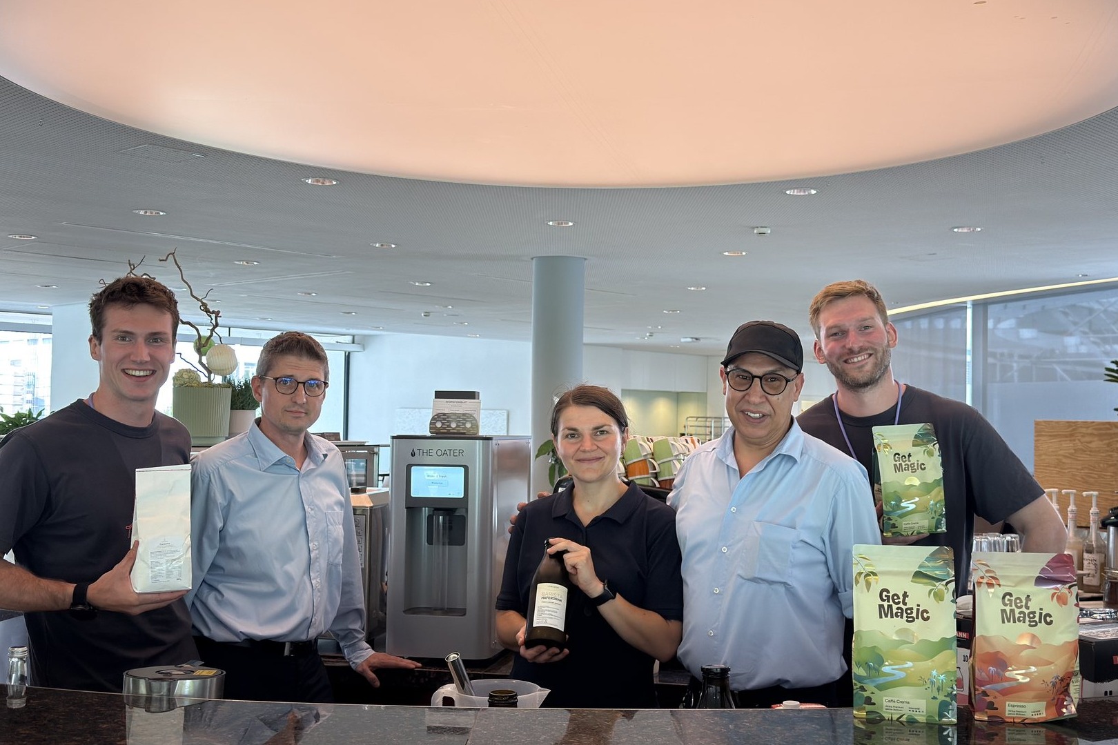 Fünf Personen stehen an einer runden Kaffeebar, dazwischen die The-Oater-Maschine, Kaffeepackungen und eine Espressomaschine