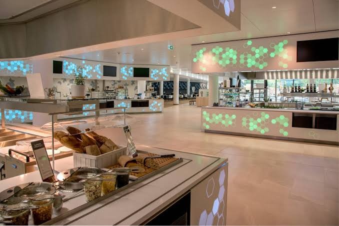 Heller Ausgabebereich einer Betriebskantine mit Buffettheke, Brotkorb, Toppings in Gläsern und hinterleuchteten Wabenmustern in Mint und Blau