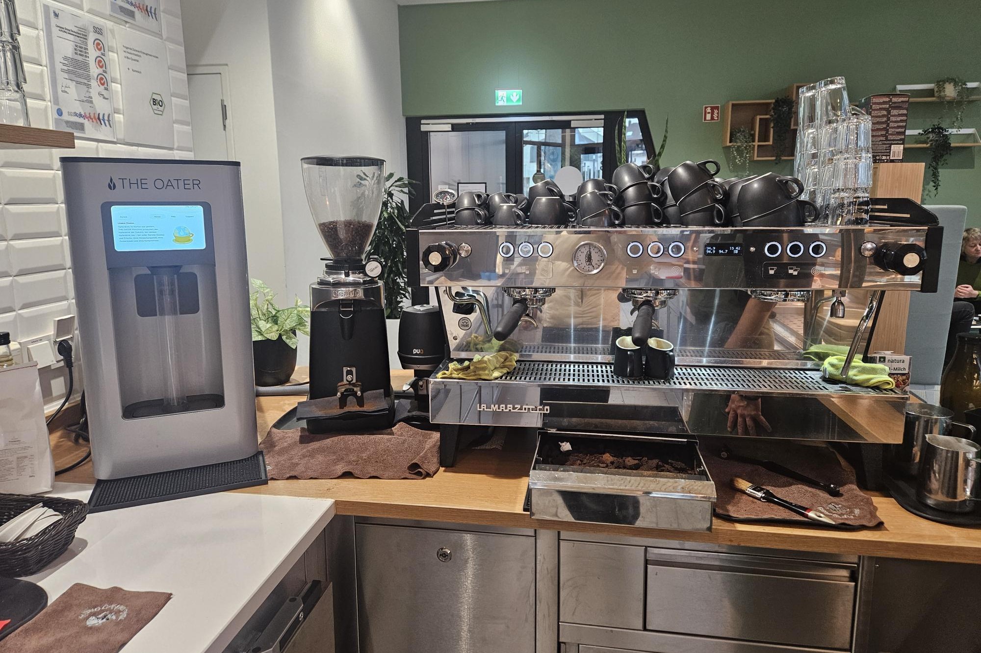 Kaffeebar mit silberner The-Oater-Maschine neben einer großen Edelstahl-Espressomaschine, schwarzen Tassen darauf und grüner Wand