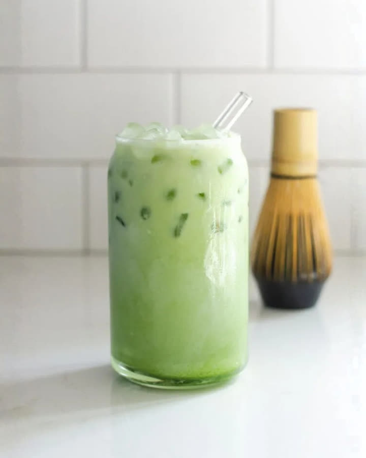 Grüner Shaken Matchagato im Glas mit Eiswürfeln und Glastrinkhalm, dahinter ein Matcha-Besen