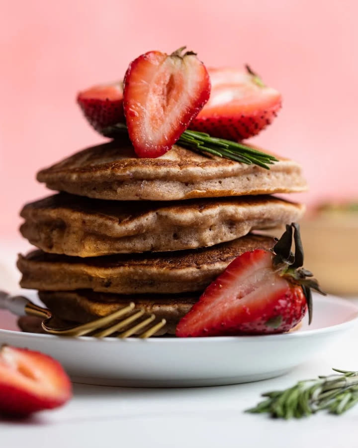 Stapel Very Berry Pancakes mit halbierten Erdbeeren und Rosmarin auf einem weißen Teller