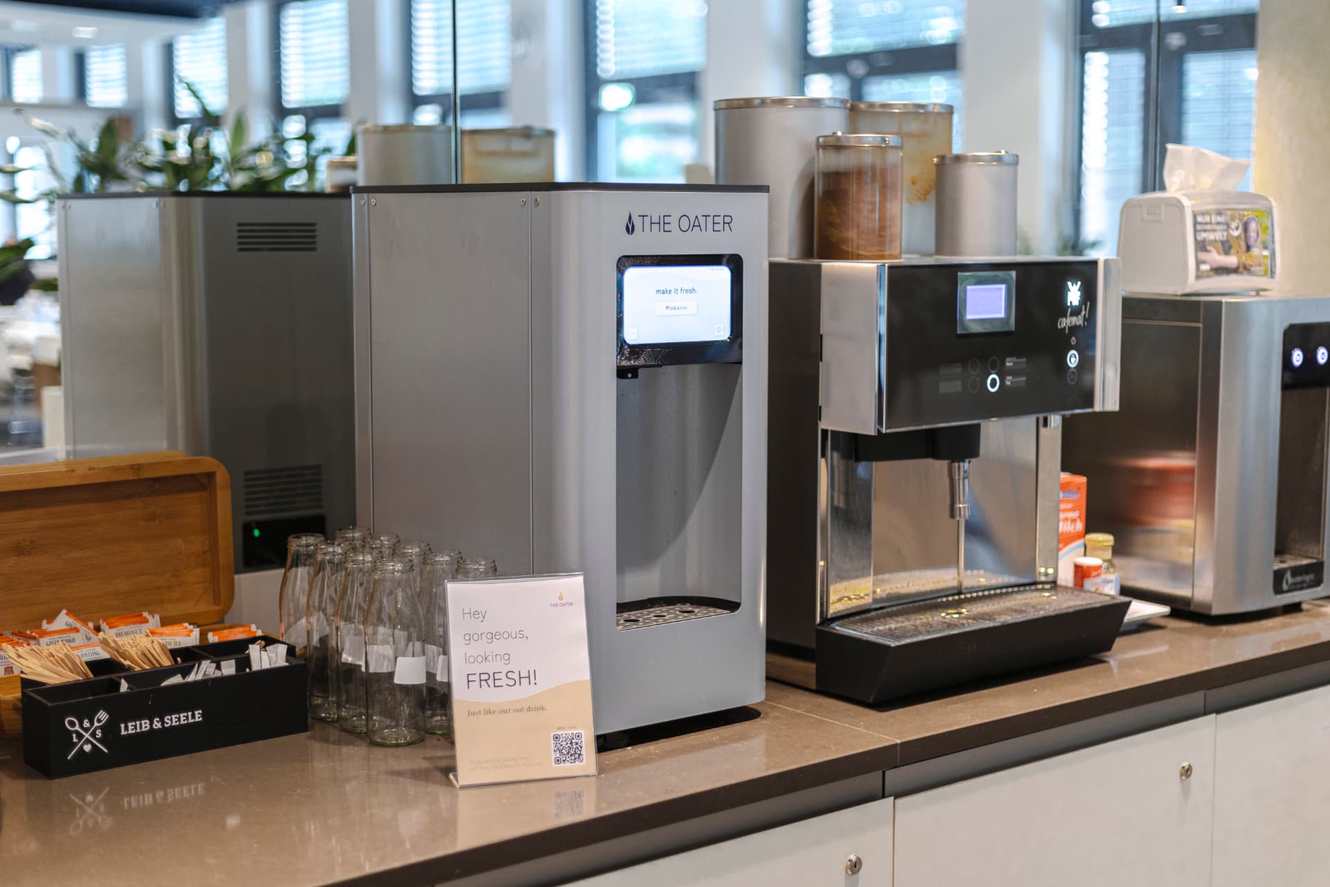 The Oater Maschine mit Display steht auf einer Theke neben einer Kaffeemaschine, davor leere Glasflaschen und ein Aufsteller, im Hintergrund eine helle Fensterfront